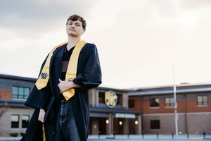 Graduate in black and gold cap and gown poses confidently outdoors in front of a school building.