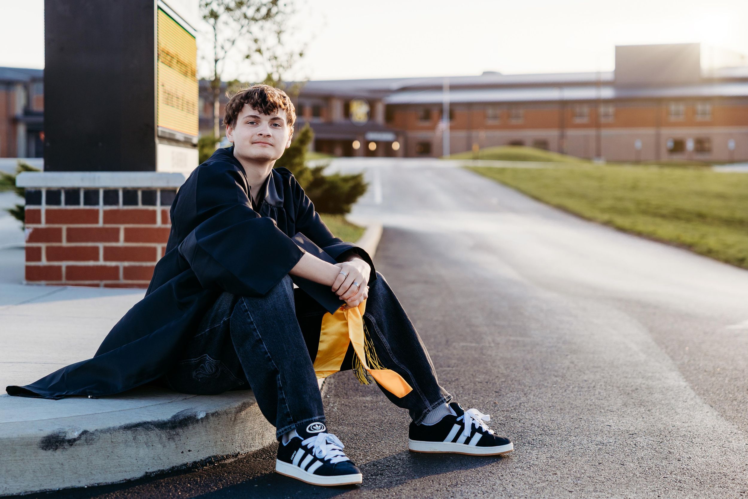 Teen boy in black outfit and Adidas sneakers sitting on steps outside a brick school building.