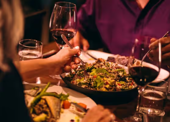 People dining at a restaurant table, clinking red wine glasses over a fresh salad and gourmet dishes in a warm, dimly lit setting.