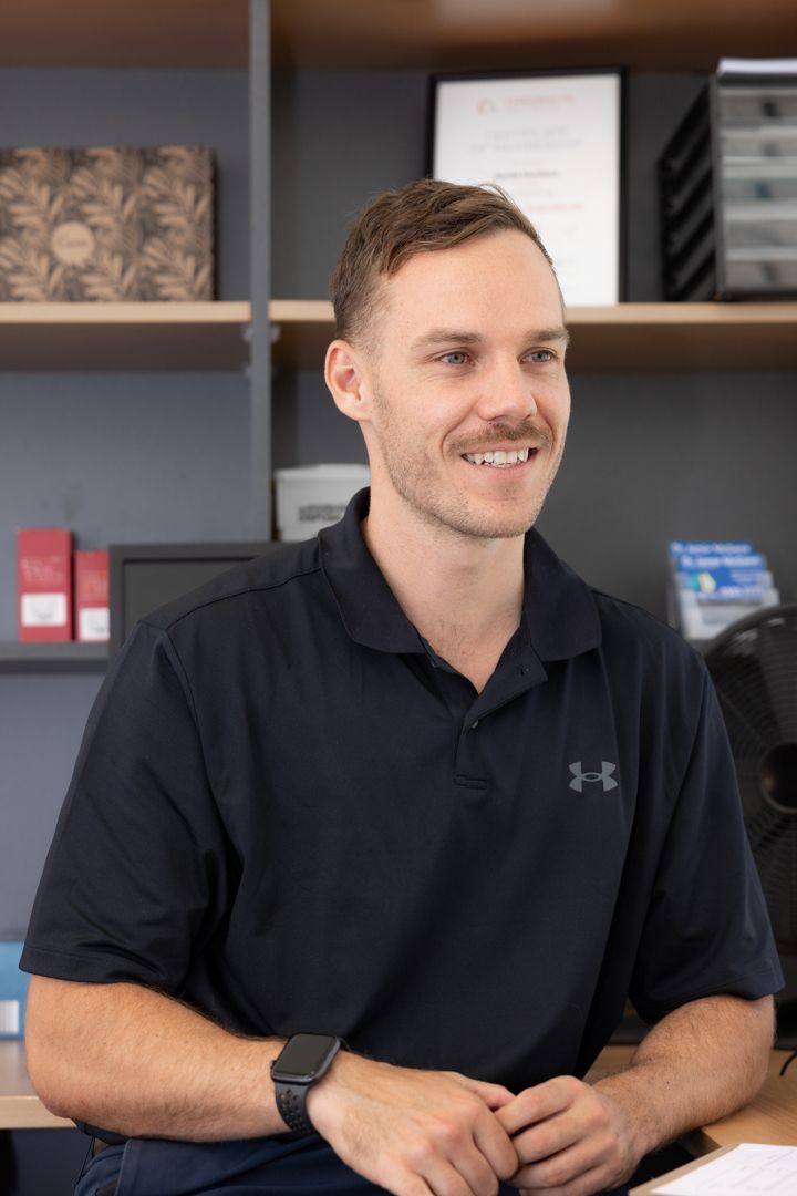 Professional in a black Under Armour polo shirt smiles warmly in an office setting with bookshelves in the background.