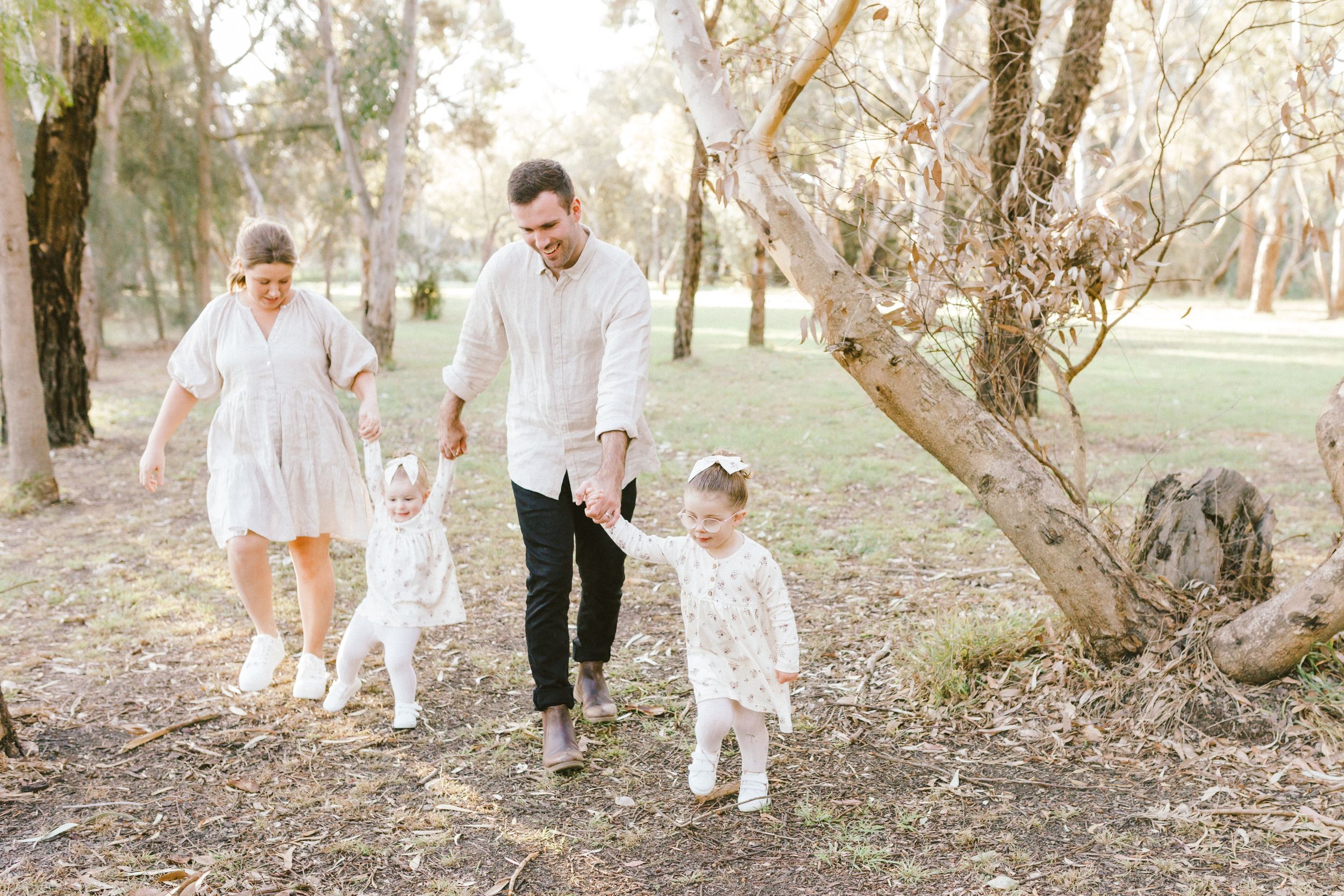 Family wearing coordinated white outfits walks together along a dreamy tree-lined path during golden hour.