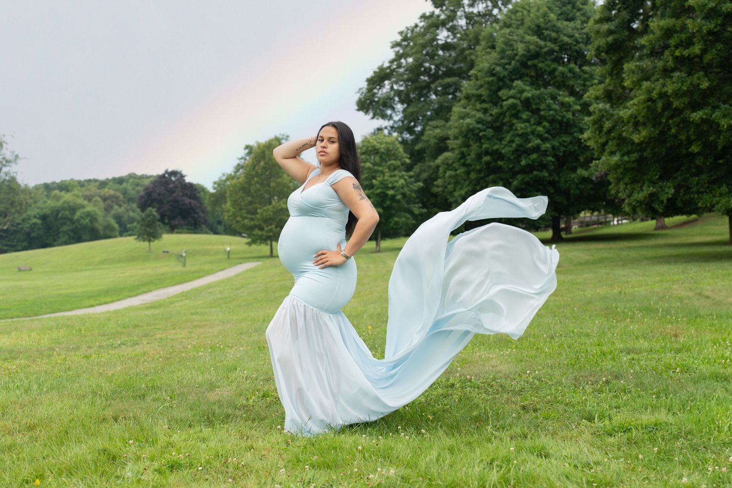 Maternity photo of client in flowing blue dress standing in field with rainbow in background.
