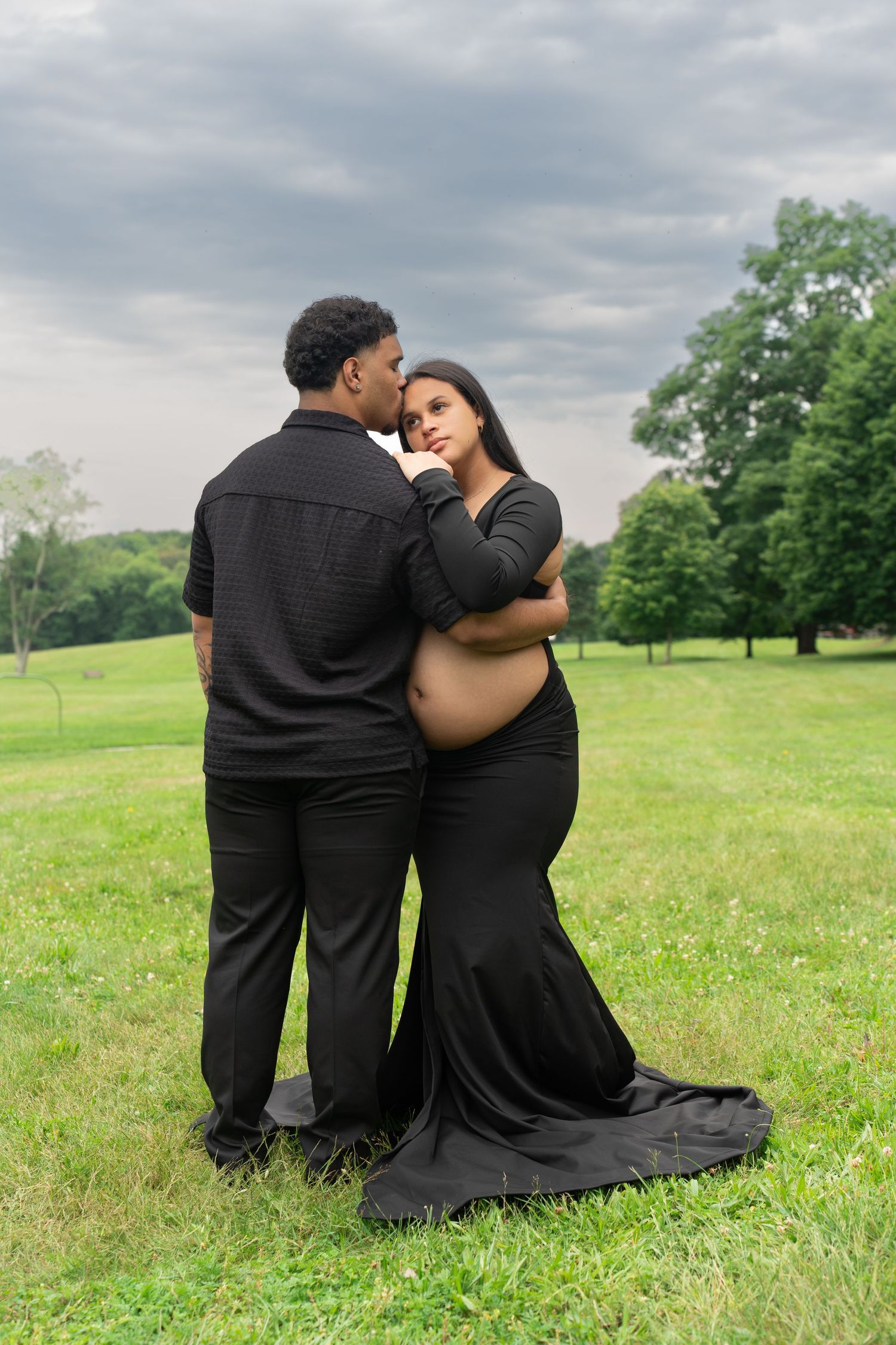 Maternity portrait of couple in black attire posing together in grassy field.