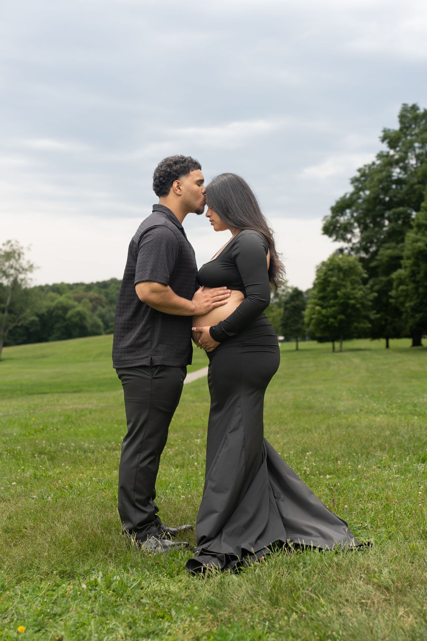 Romantic maternity portrait of couple in black clothing embracing in park setting.