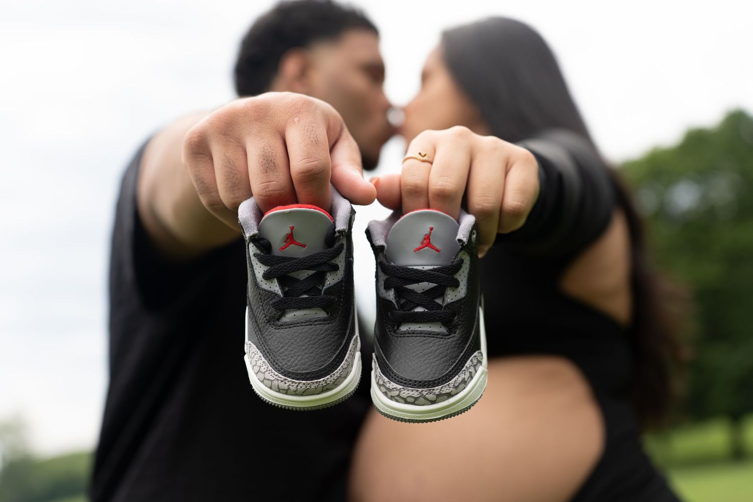 Baby shoes held up in foreground with couple embracing in background at park.