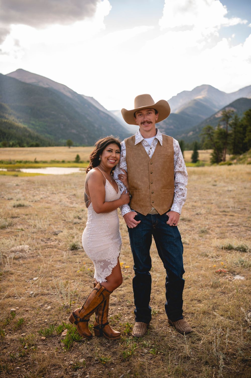 A couple poses in a western setting with mountains and fields in the background wearing wedding attire and cowboy boots.