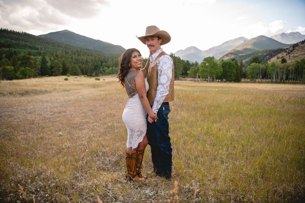 A romantic couple embraces in a golden meadow with majestic mountains and forest in the background on a summer evening.