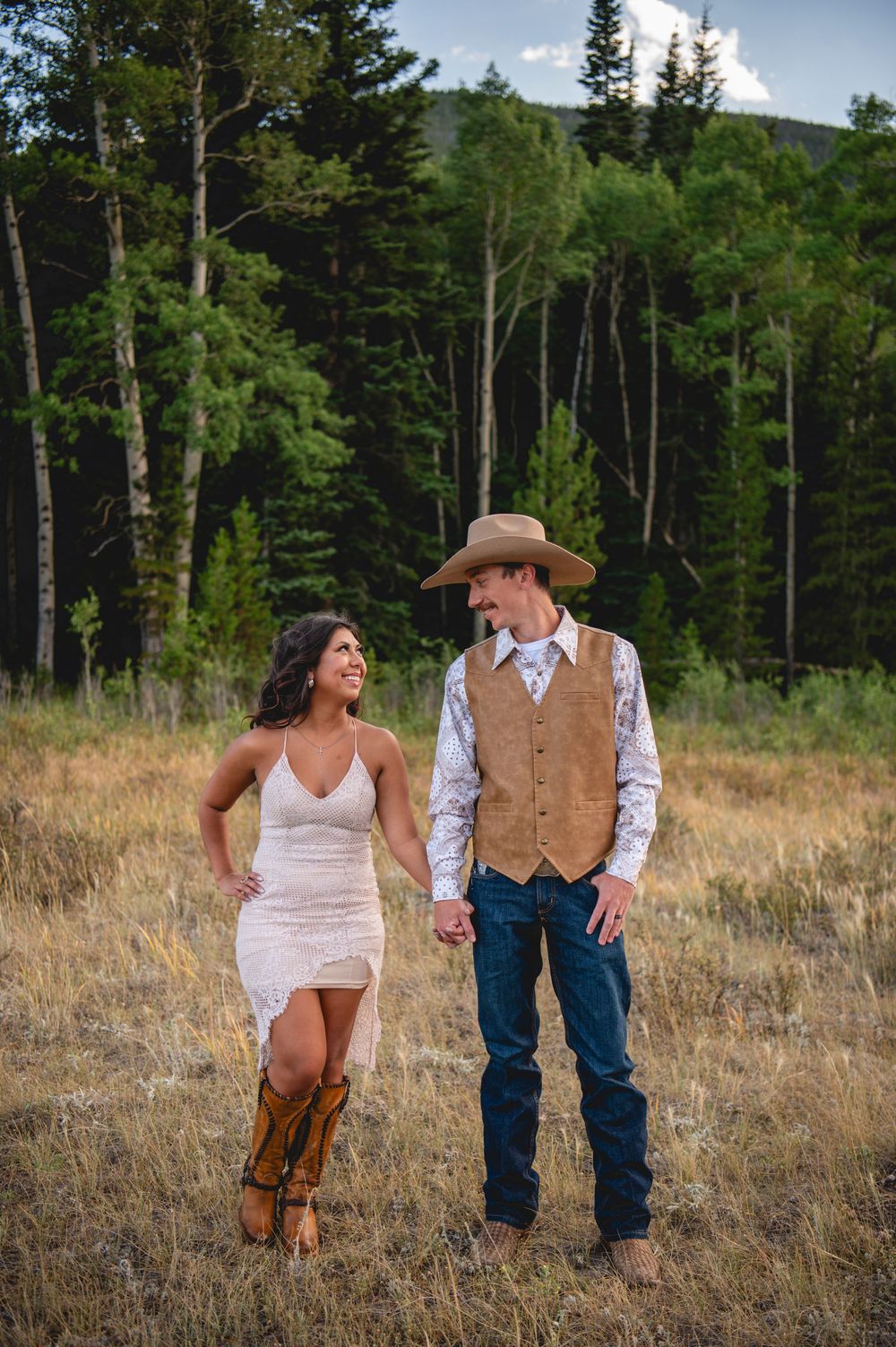 A couple in western attire stands together in a mountain meadow with tall pine trees in the background.
