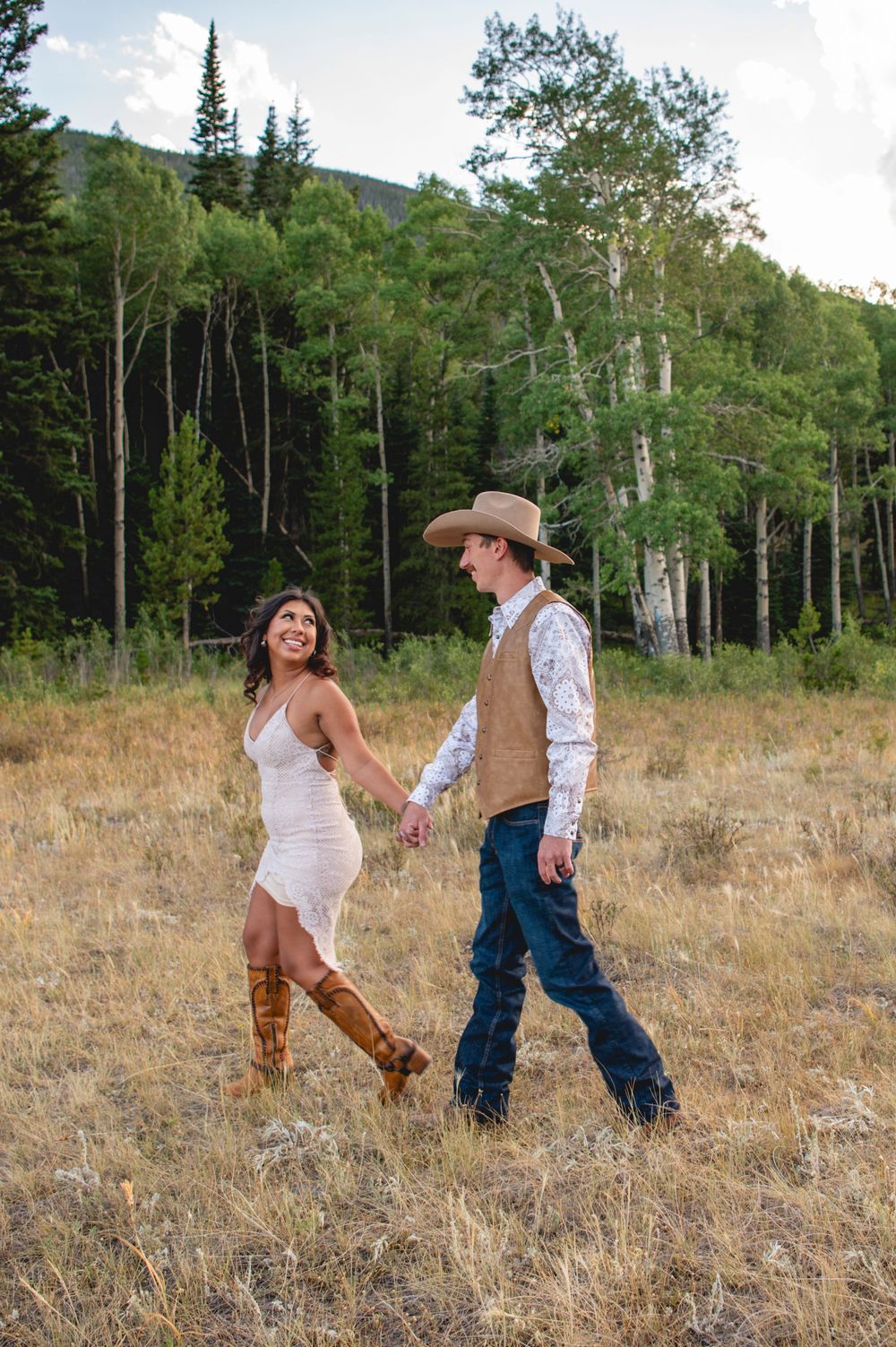 A playful couple dances together in a grassy meadow surrounded by aspen and evergreen trees at sunset.