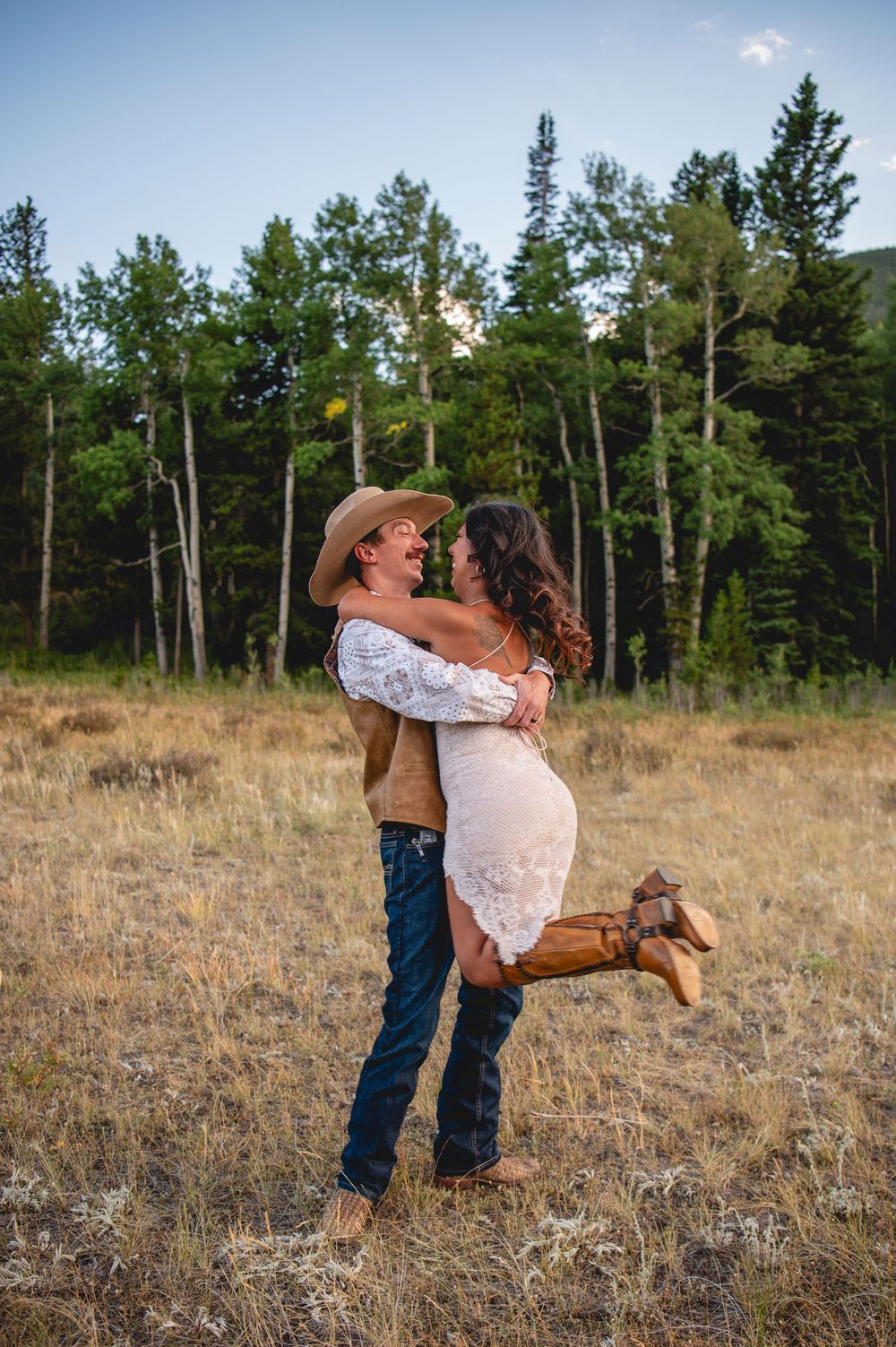 A couple embracing in a romantic dance pose in a rustic field with evergreen trees in the background at sunset.