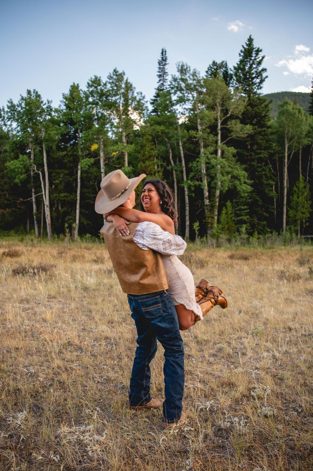 A romantic outdoor moment captured in a field with tall pine trees and mountains in the background.