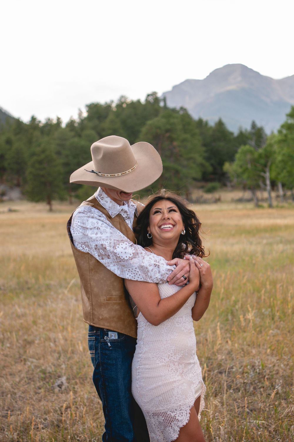 A romantic outdoor portrait session showing a couple embracing in a scenic mountain field at sunset.