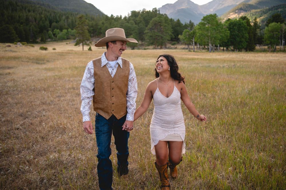 Two people holding hands while walking through a rustic golden meadow with mountains in the background at sunset.