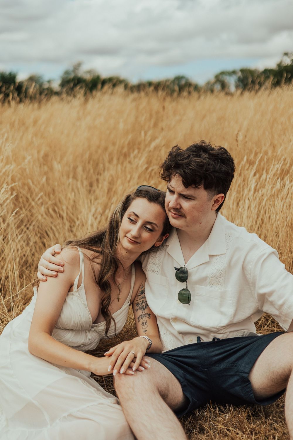 A couple sits together in a golden wheat field, the woman leaning on the man's shoulder in a tender embrace.