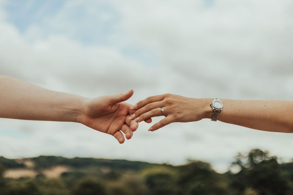 Two hands reaching toward each other against a cloudy sky, one wearing a silver watch, symbolizing connection and love.