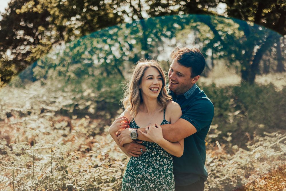 A romantic couple embraces and laughs while standing outdoors during an engagement photoshoot at sunset.