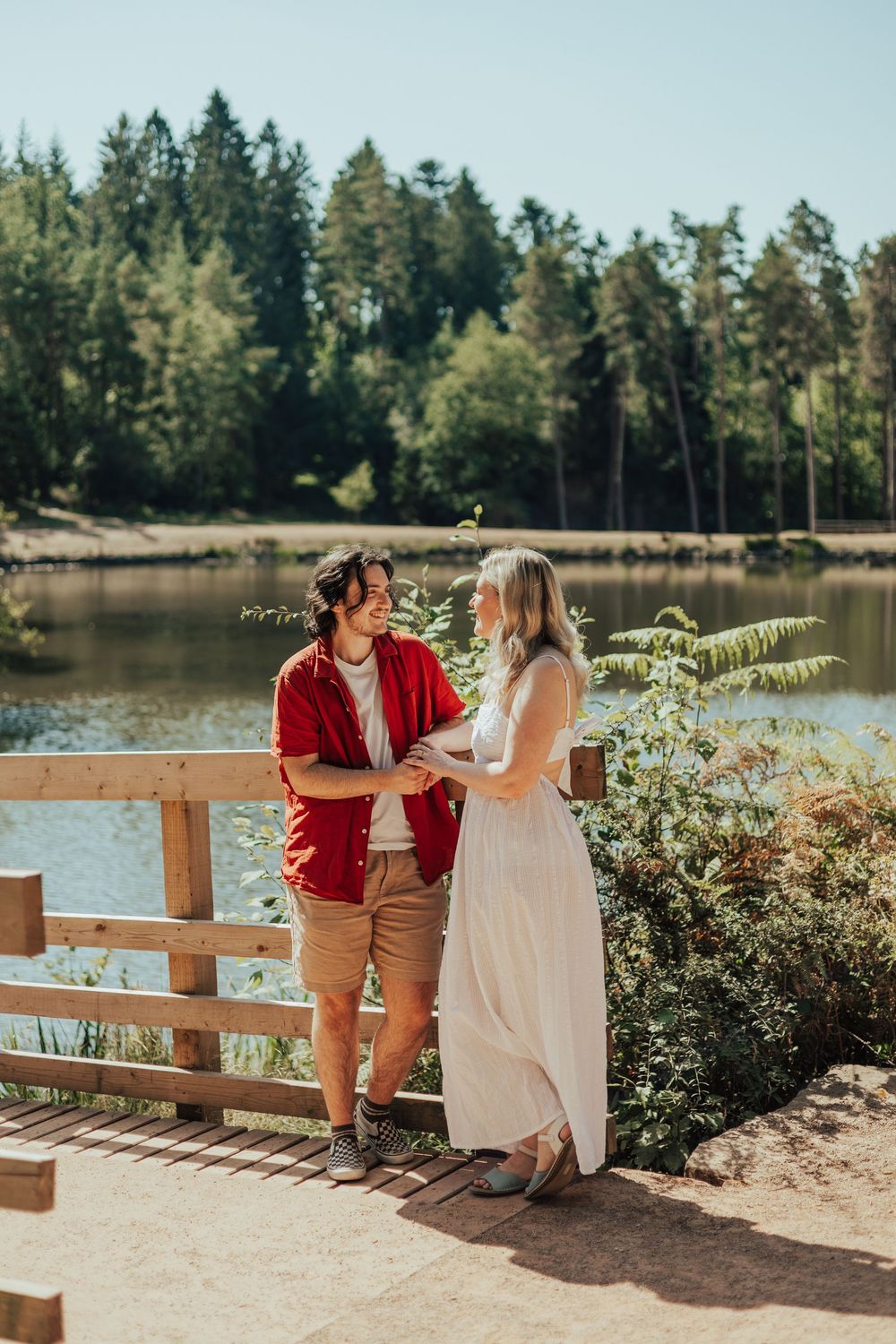 Couple holding hands by a wooden fence near a lake, surrounded by lush green forest on a sunny day.