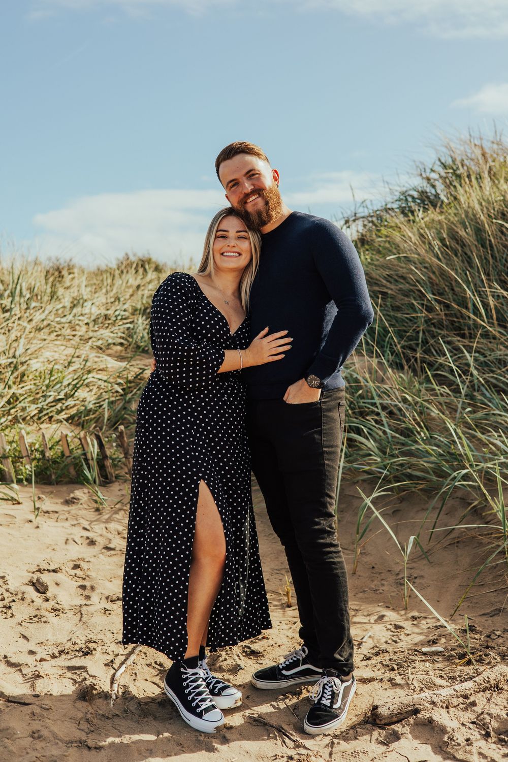 A couple shares intimate moments against a backdrop of beach dunes and tall grass on a sunny day.