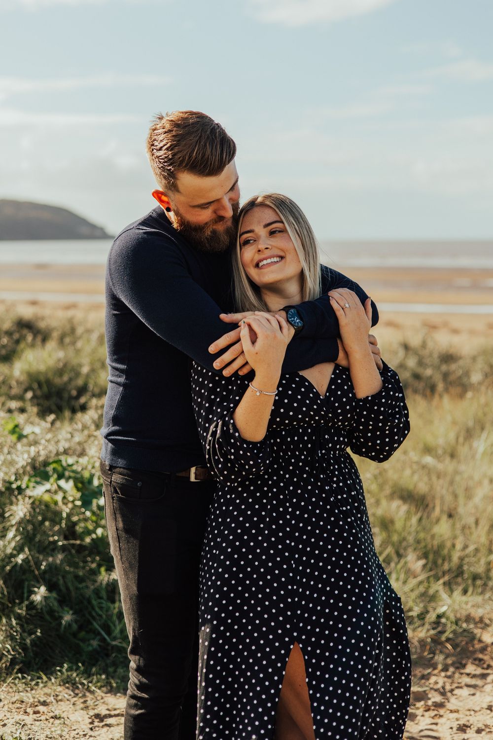 A couple shares a tender moment outdoors in a polka dot dress and dark clothing against a natural landscape backdrop.