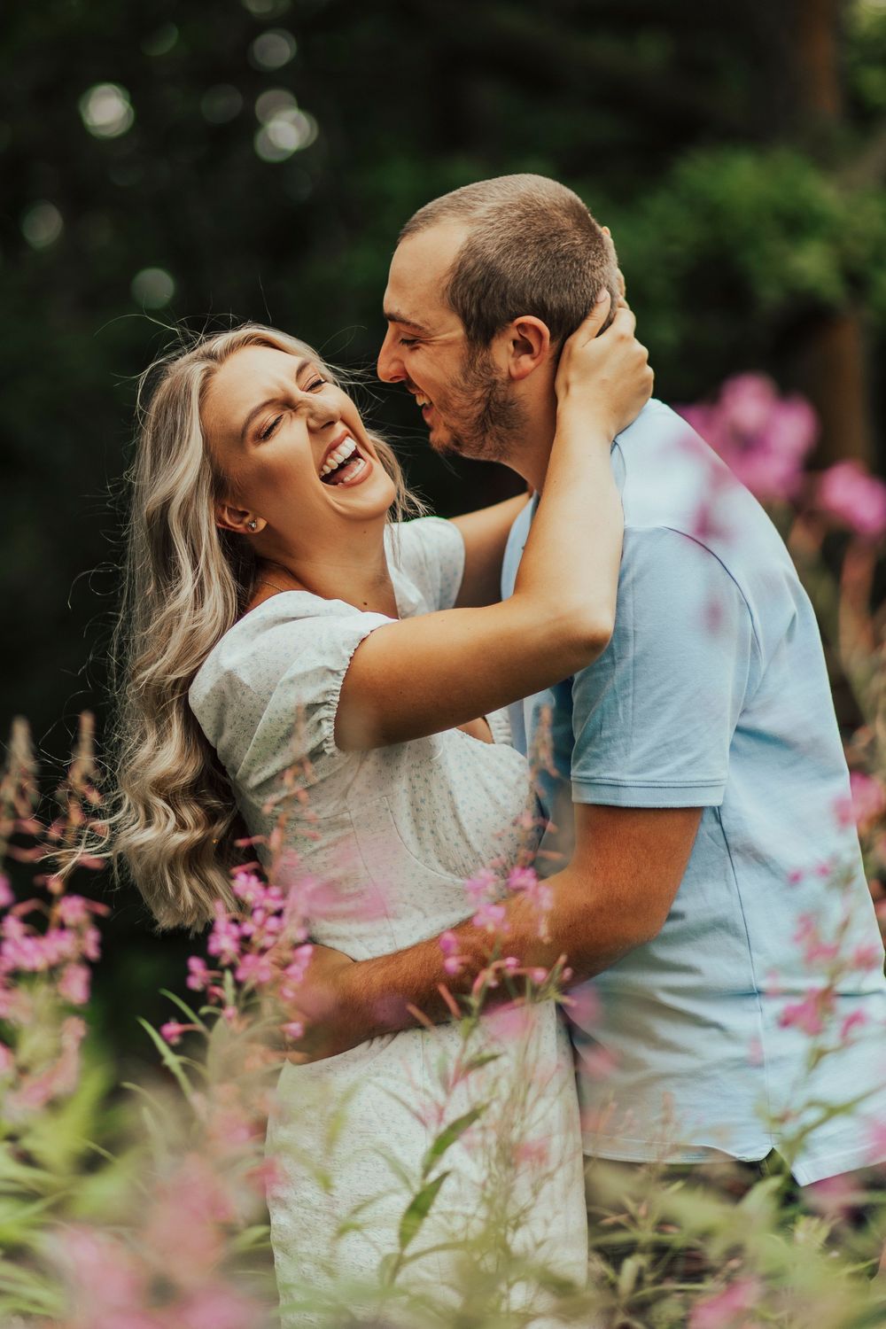 A couple embraces and laughs joyfully in a garden with pink flowers in the foreground.