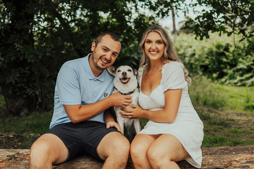 A happy couple sits outdoors with their small white dog on a sunny day in a natural setting.