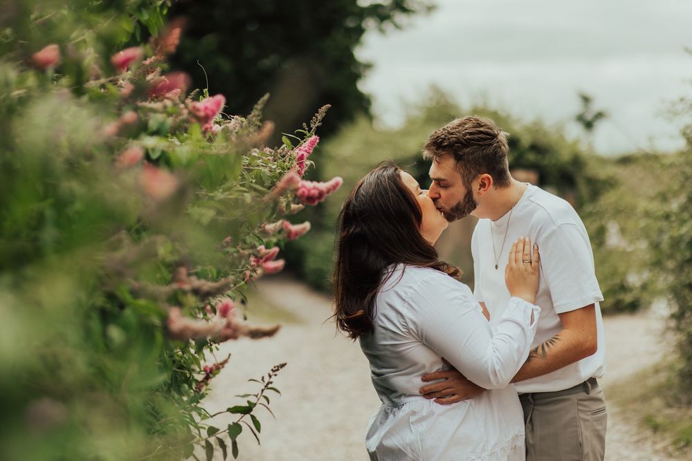 Couple embraces tenderly in a lush garden setting with pink flowers and natural greenery in the background.