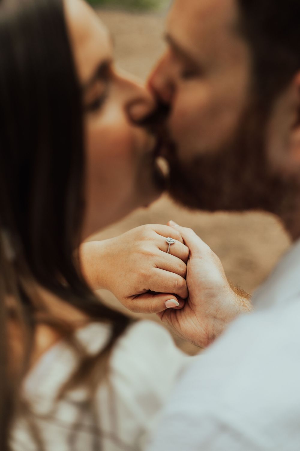 Couple sharing a romantic kiss while holding hands, showcasing an engagement ring in a soft-focus close-up photo.