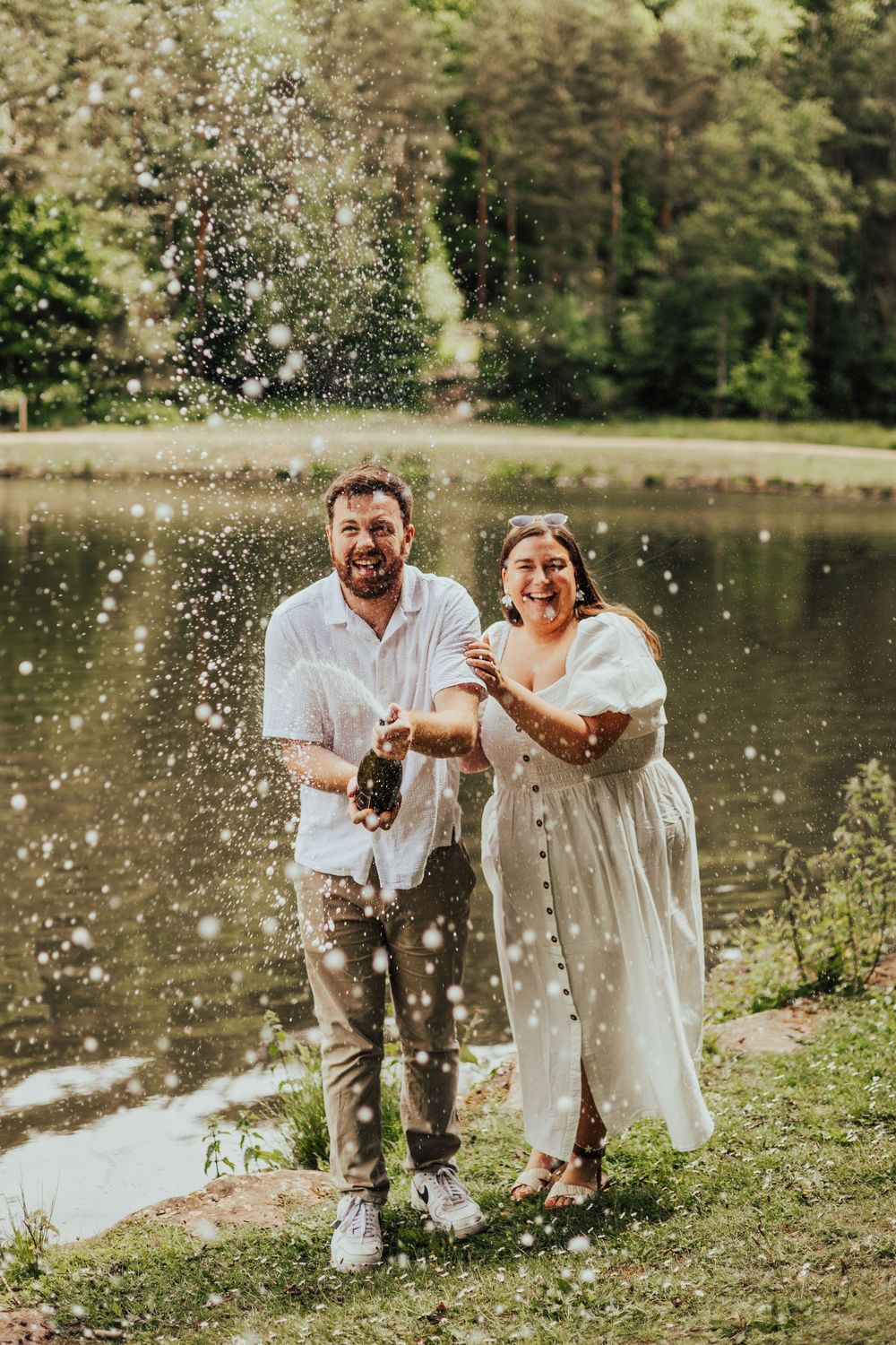 Joyful couple popping champagne bottle by a serene lake surrounded by lush green trees on a sunny day.