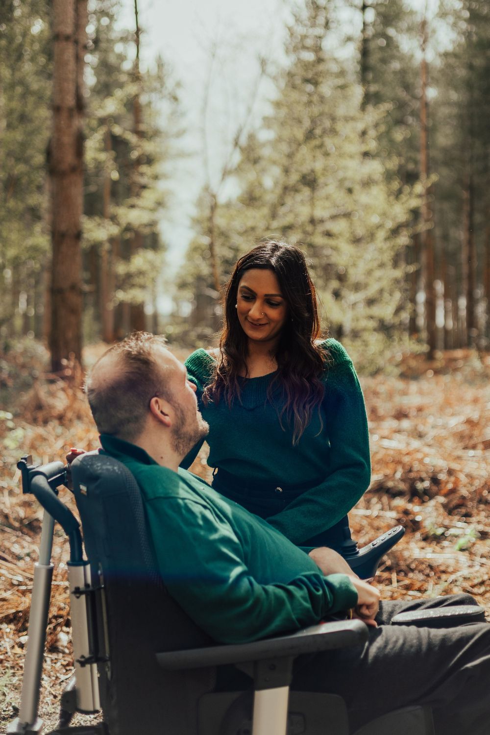 A woman in teal smiles at a man in a wheelchair surrounded by tall trees in a peaceful forest setting.