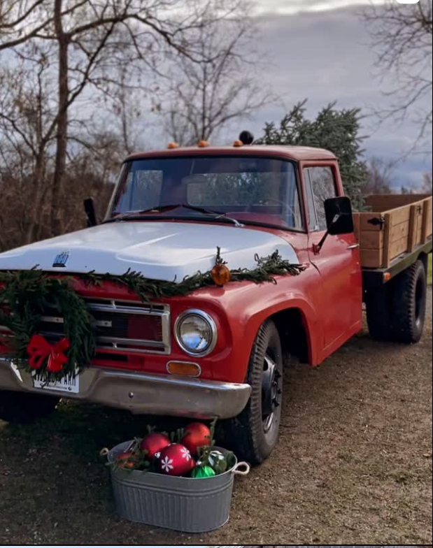 Red vintage truck decorated with Christmas ornaments and a wreath sits parked outdoors on a cloudy day.