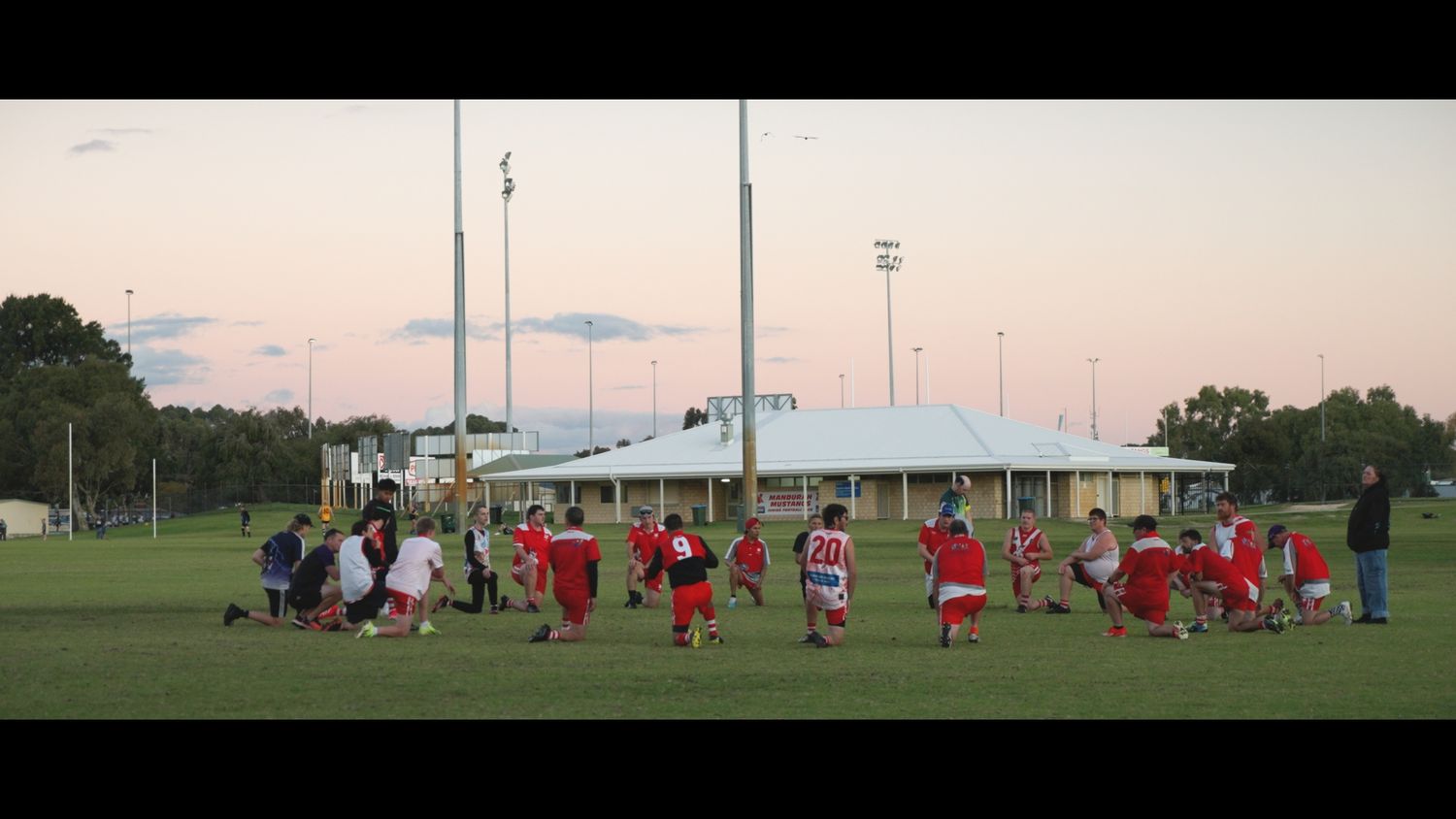 Youth sports team and spectators gather on a grass field at sunset with field lights and a covered pavilion in background.