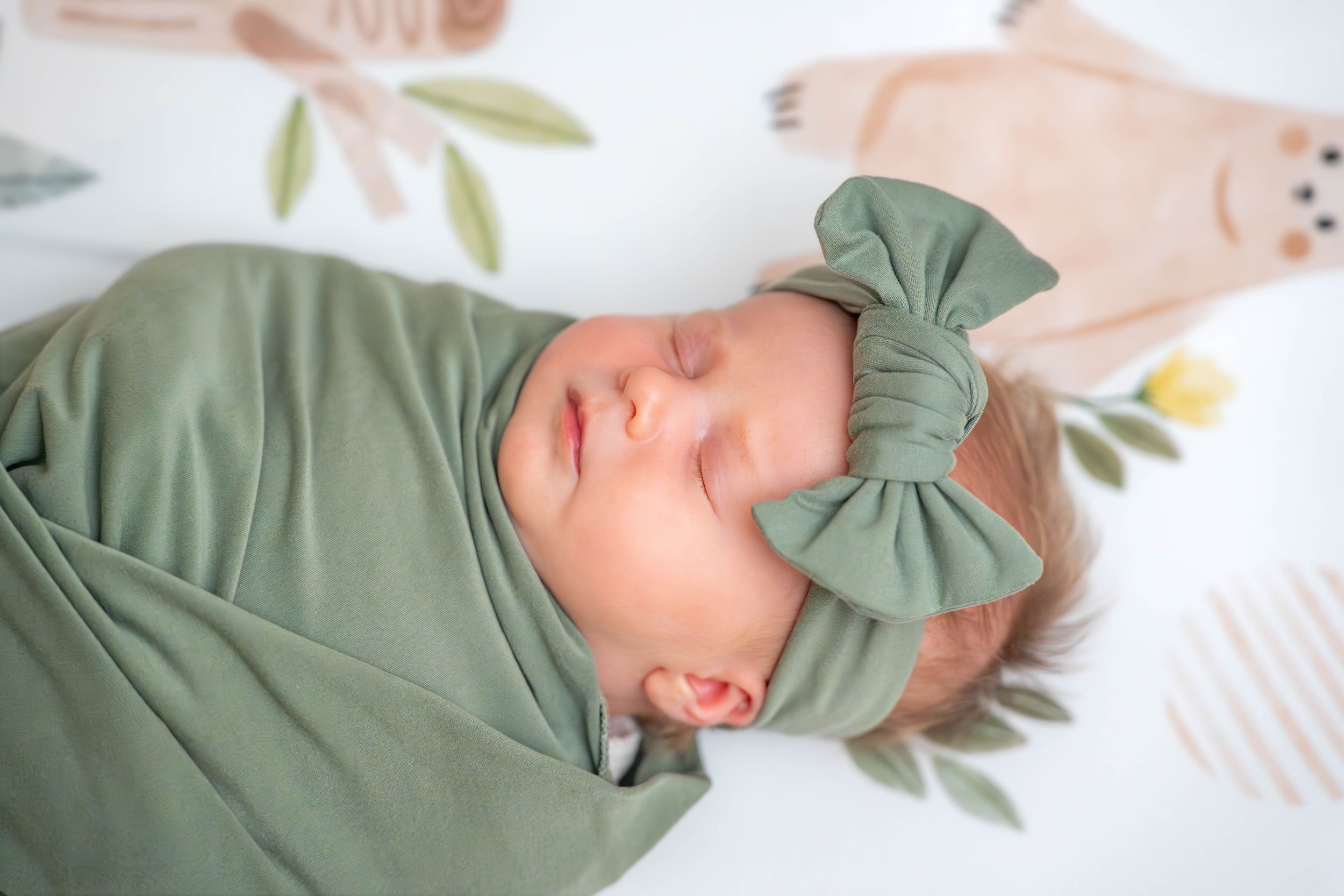 Sleeping newborn wearing sage green outfit with bow accessory on white bedding. Quad Cities Newborn Photography