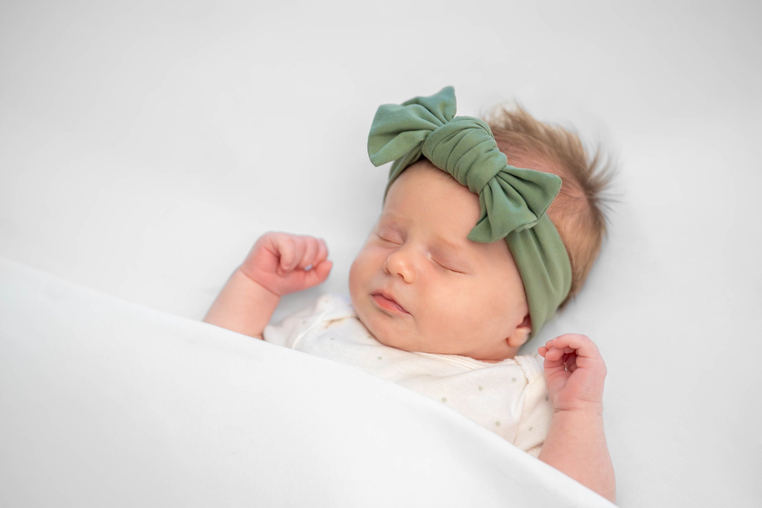 A sleeping baby wearing a mint green bow headband rests peacefully on a soft white blanket. Quad Cities Newborn Photography
