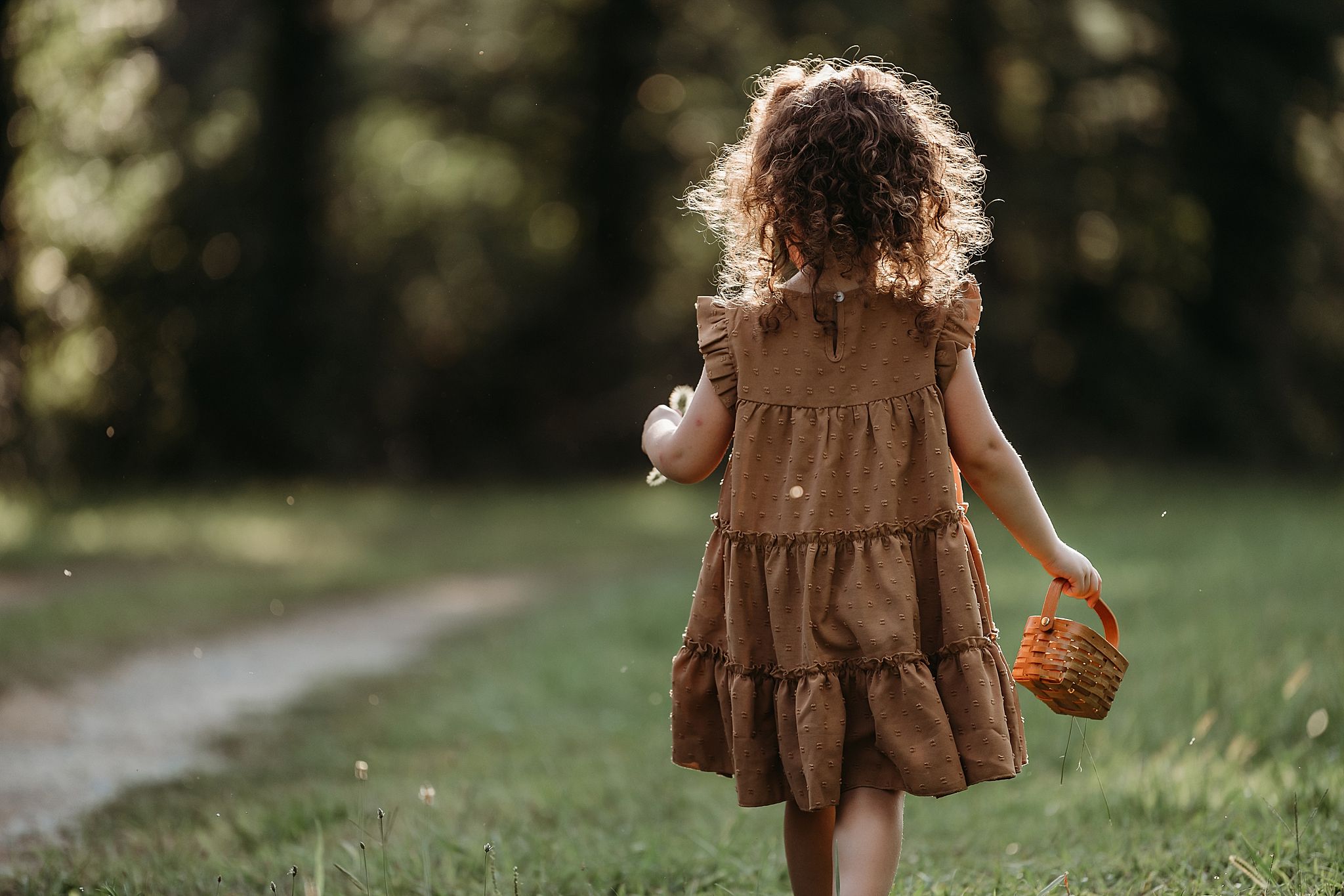 A child in a brown dress with curly hair walks down a dirt path carrying a basket on a sunny day.