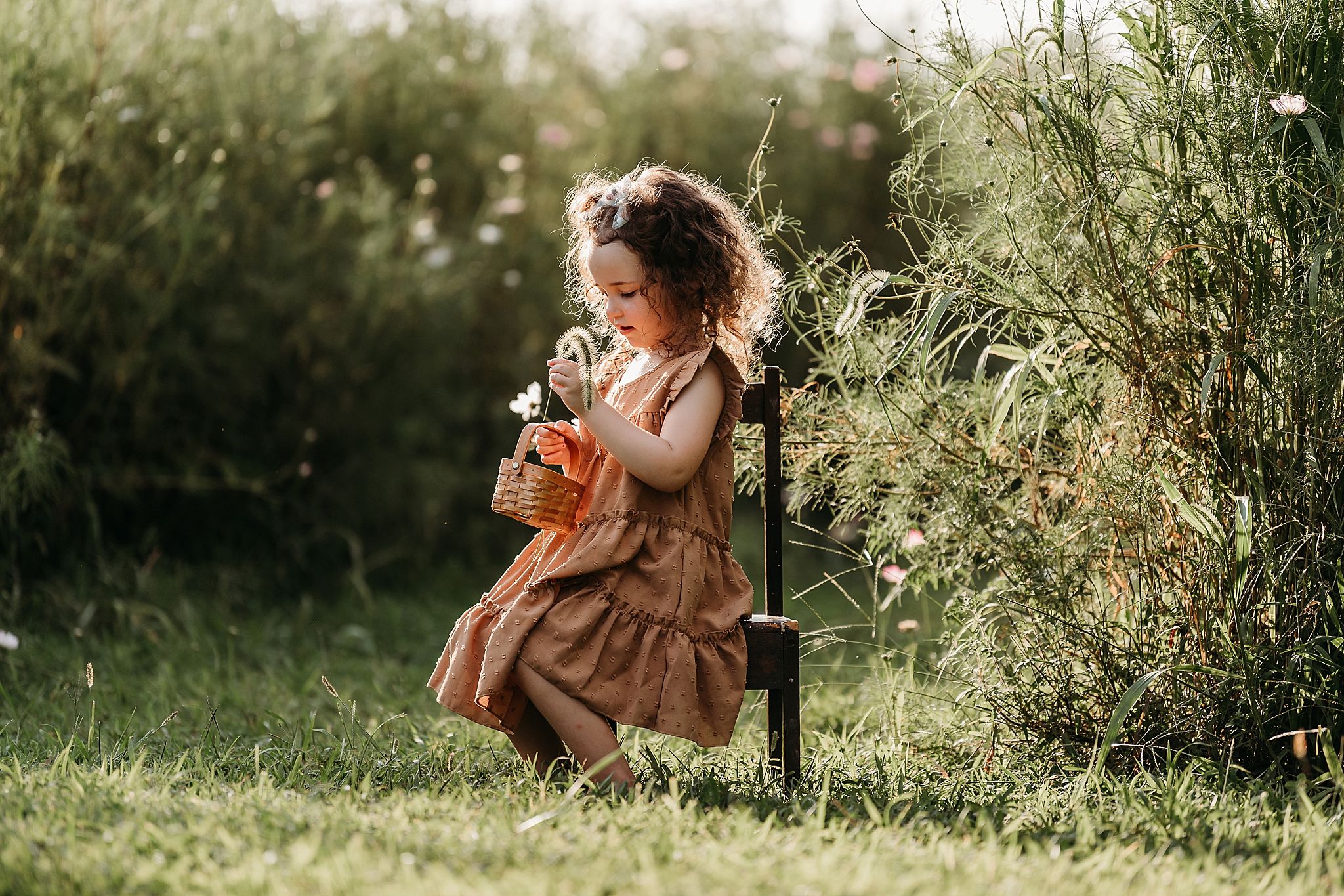 A child in a brown dress kneels in lush grass on a sunny day while smelling flowers.