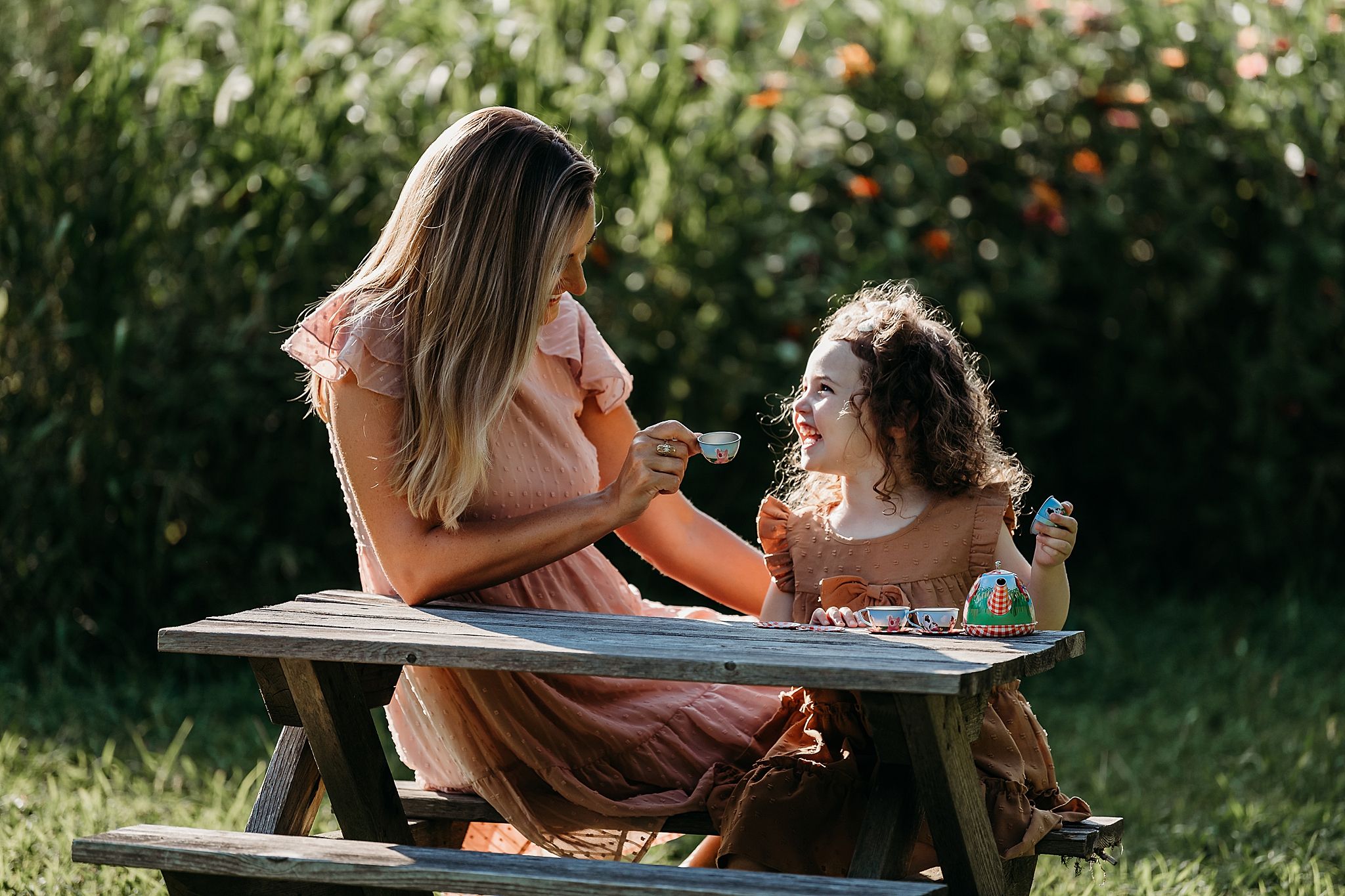 Two people sit together at a wooden picnic table in a sunny garden while sharing a joyful moment.