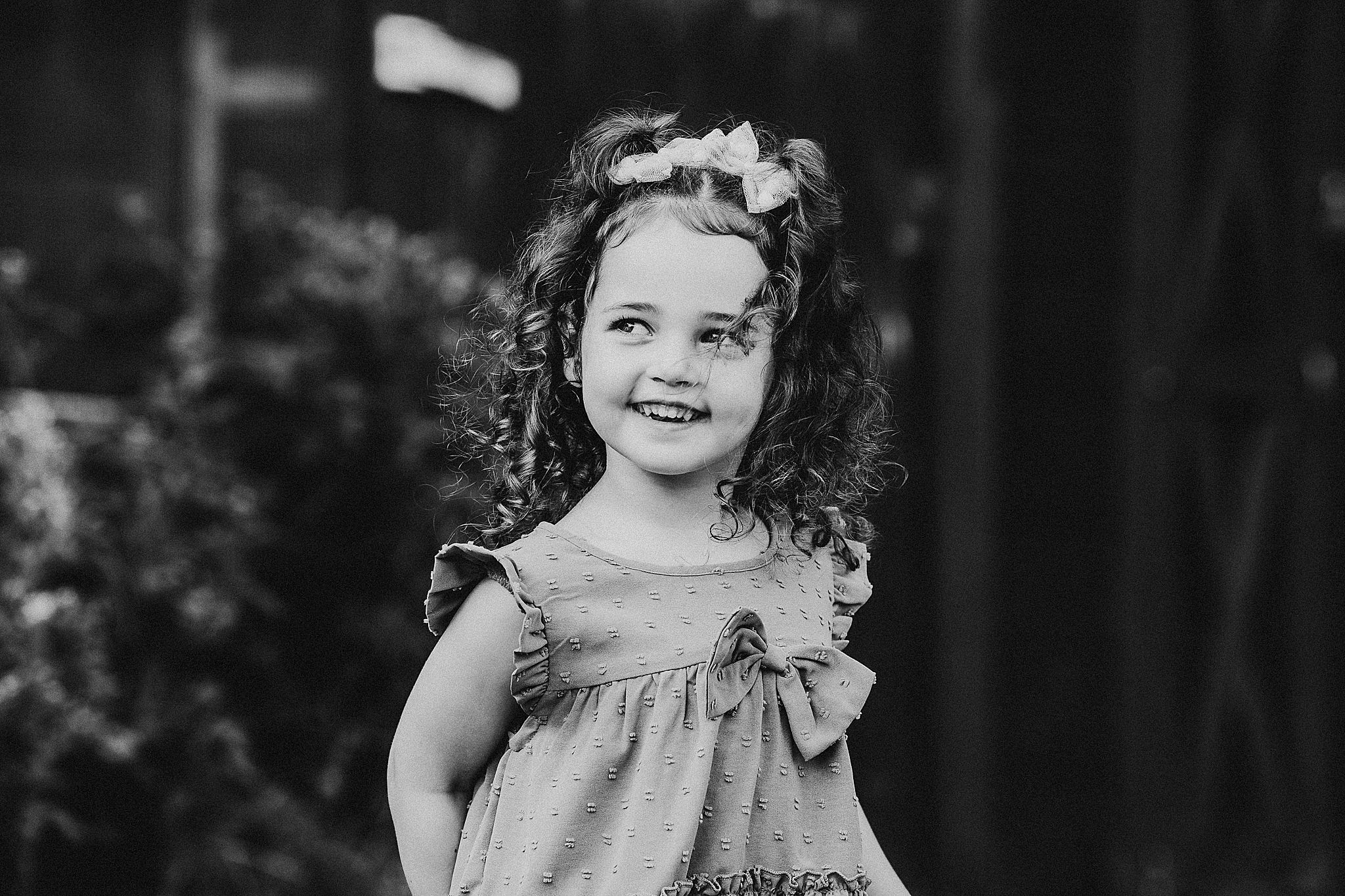 Smiling child in black and white portrait photo wearing a floral dress against dark backdrop.