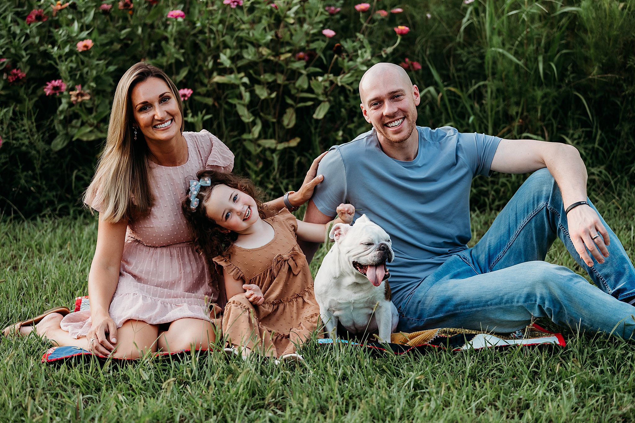 A joyful family and their white dog sit together on green grass in front of flowering bushes on a sunny day.