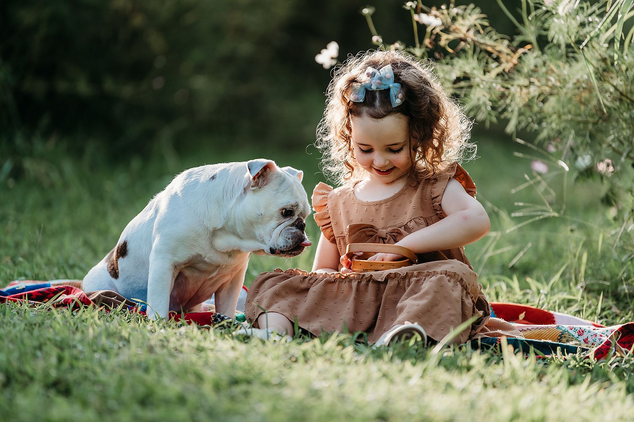 A spotted white dog and a child in a brown dress share a tender moment while sitting on grass during a picnic.