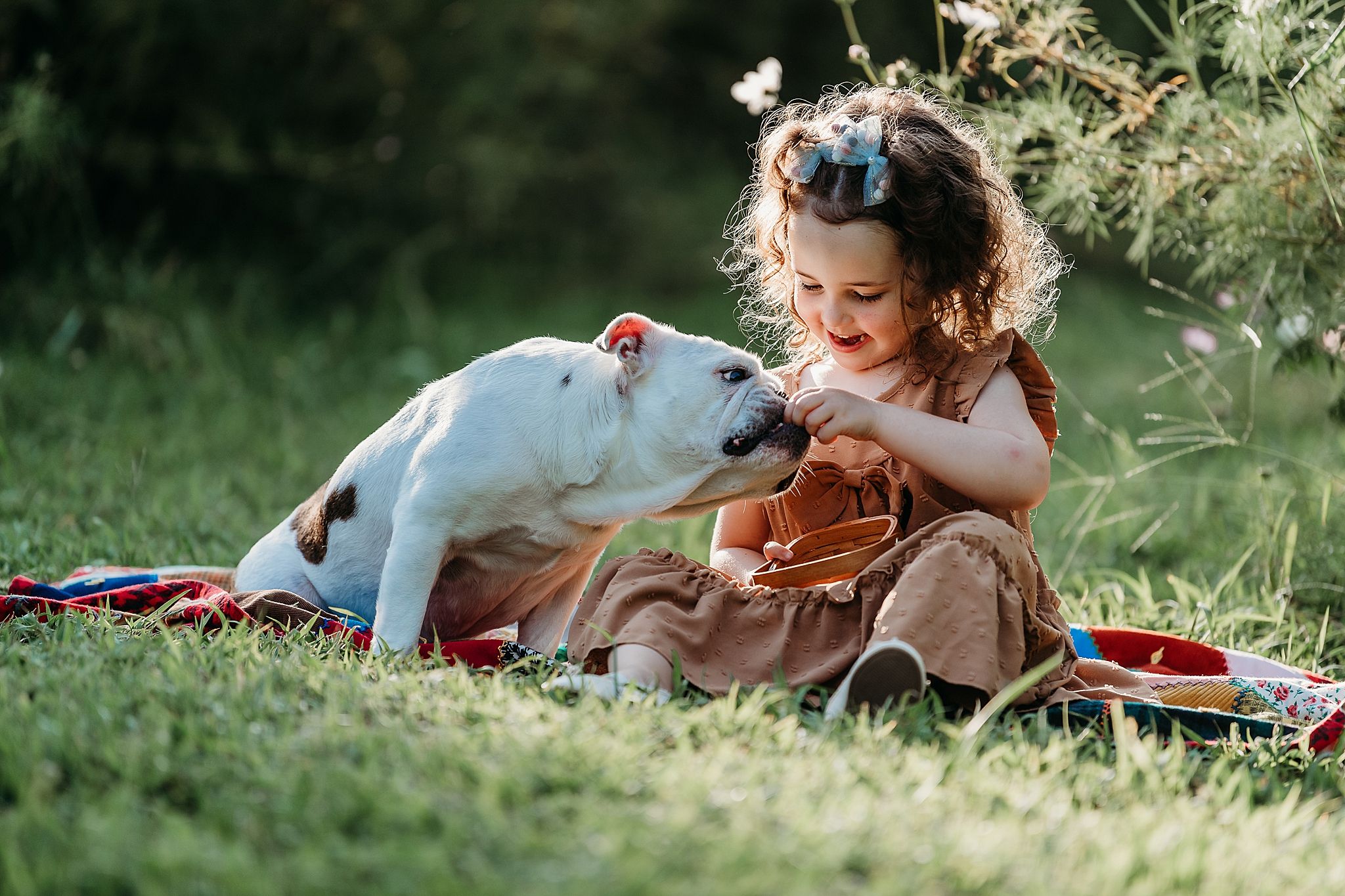 A child in a brown dress sits on grass petting a white dog during a sunny summer picnic.