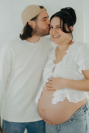 A romantic maternity photoshoot features an expecting couple in white shirts and jeans embracing against a bright background.
