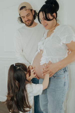 Family expecting new baby poses together in white room with young child touching pregnant mother's belly.