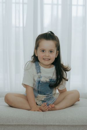 A child in denim overalls sits in a bright white room with soft sheer curtains in the background.