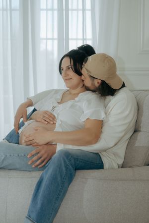 A couple in casual attire resting together on a light gray couch in a bright sunlit room.