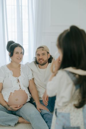 An intimate indoor maternity session shows a couple in casual white outfits sharing tender moments by a window.