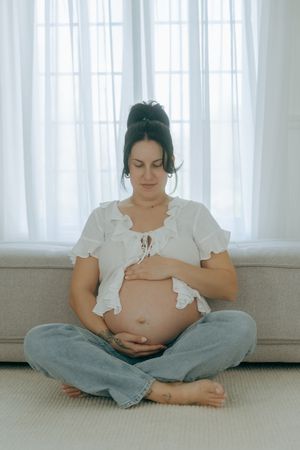 A pregnant woman in casual clothing sits cross-legged by a window in contemplative maternity poses.