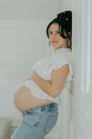 A pregnant woman in a white ruffle top and jeans poses against white walls in a bright, minimalist setting.
