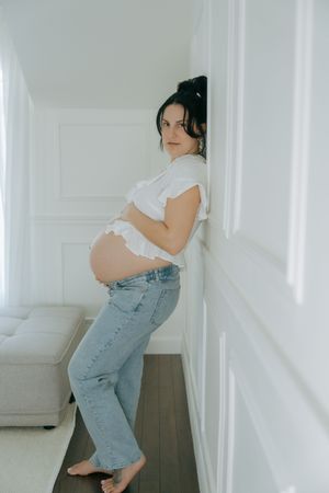 An expectant person in light top and jeans poses against a white wall in a minimalist indoor setting.