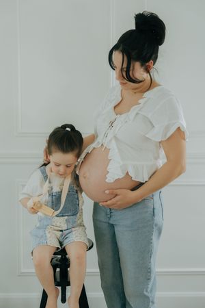 A parent in jeans and white top embraces their toddler wearing denim overalls in a minimalist white room photoshoot.