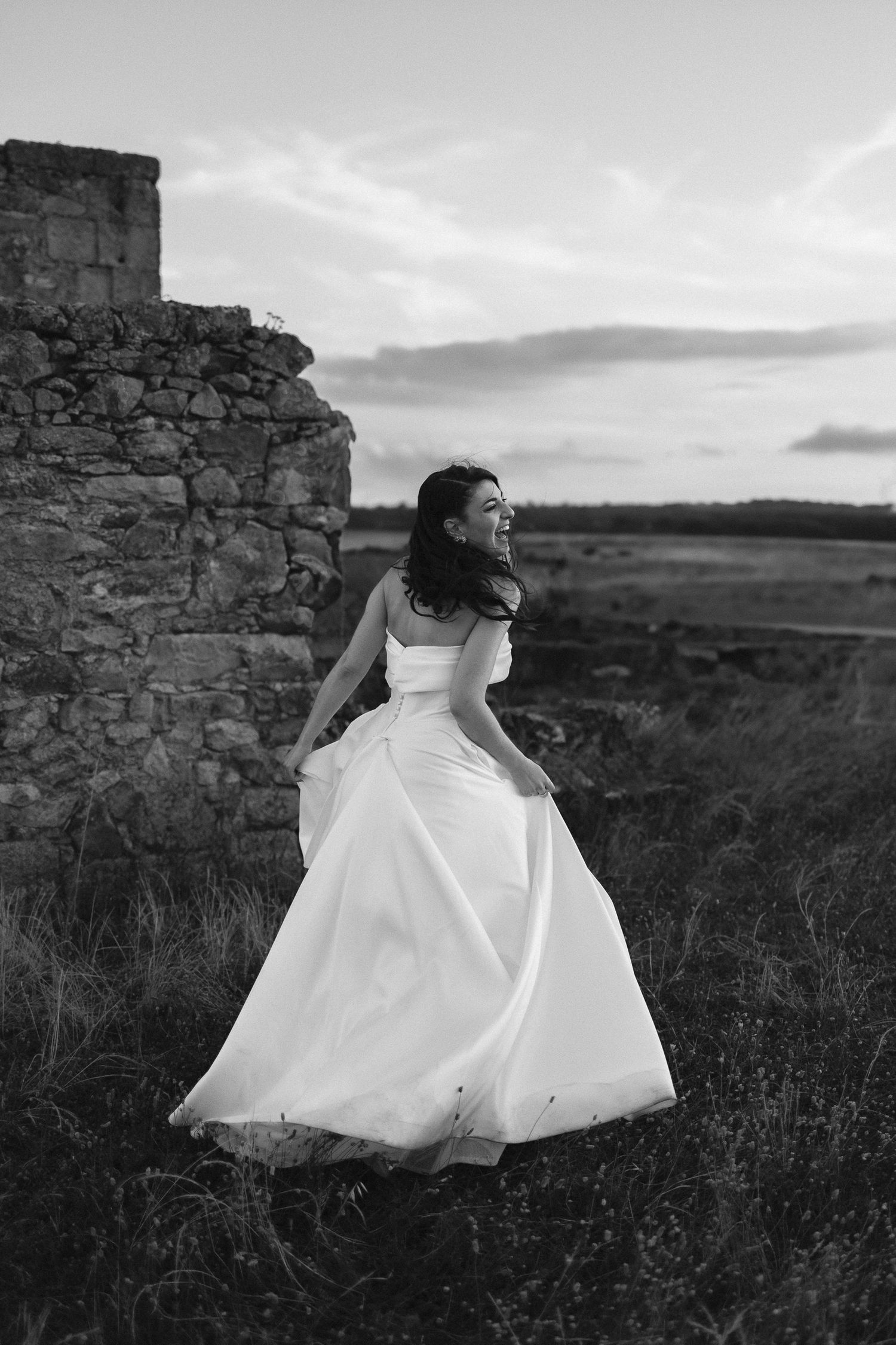 A figure in a flowing white wedding dress stands near stone ruins at sunset in black and white photography.
